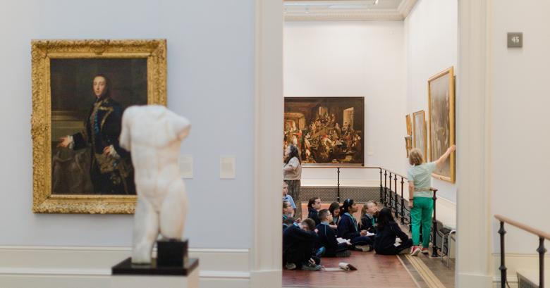 Through an open doorway in the Gallery we can see a group of young children in school uniforms sitting on the floor gazing up at a painting that a guide is showing them.