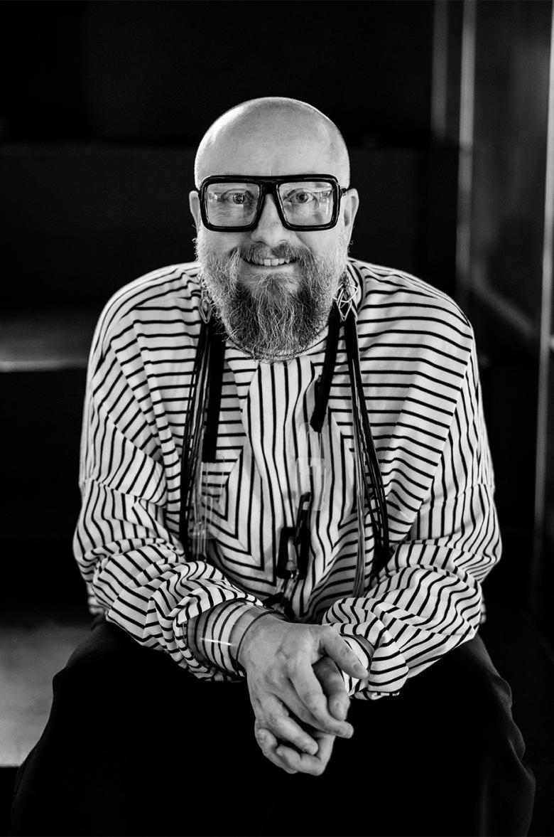 Black and white photo of a man who is bald, wearing thick rimmed black glasses, a long beard and a black and white striped top