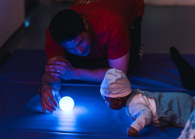 A man holds a bright blue round light on the floor in front of a baby who is crawling towards it.