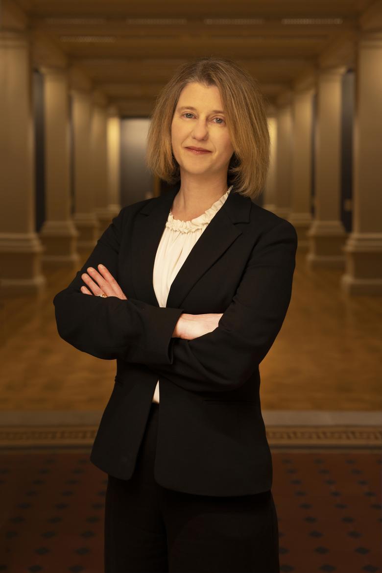 The director of the National Gallery of Ireland stands in three quarter length, arms folded, wearing a black suit over a white shirt, Behind her, the columns of a gallery are softly lit.