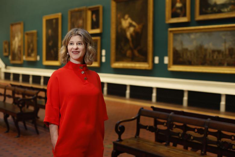 A woman in a red dress stands in a Gallery hung with large oil paintings. 
