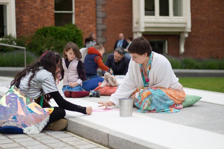 A group of people sit outside on the ground, drawing in crayons and pastels