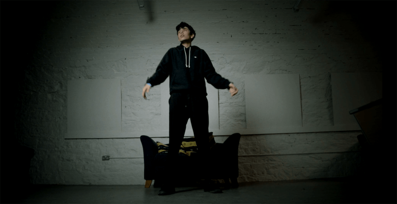 A film still of a person wearing black clothes dancing in front of a black armchair in a room with white walls
