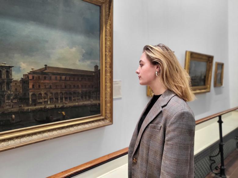 A woman stands in front of a large oil painting, looking closely at it.