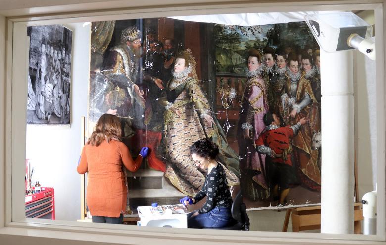 View through a window into a conservation studio where two women work on a large oil painting