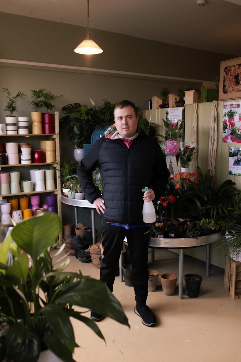A man in a black puffer jacket and tracksuit stands with one hand on his hip in a room filled with pot plants. In his other hand, a spray gun filled with water. 
