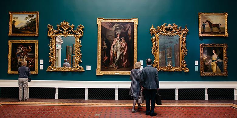 A man and a woman look at art in a grand room filled with painting and antiques.
