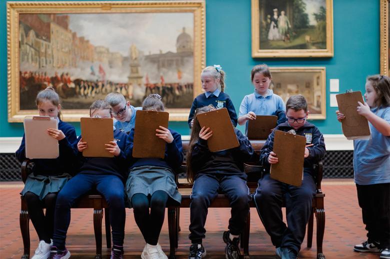 A group of school children in uniform draw on clipboards in a grand gallery filled with paintings.
