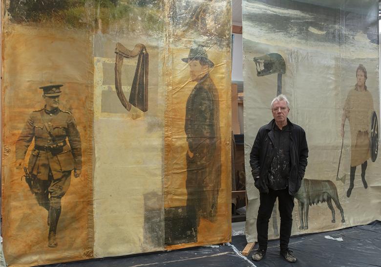 Artist Hughie O'Donoghue standing in his studio in front of two large artworks depicting Michael Collins and Wuffa.
