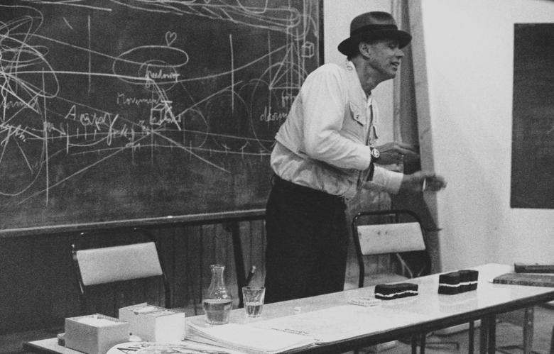 A black and white photograph of Joseph Beuys delivering a lecture at the Crawford Art Gallery. He is animated, in the process of speaking. Behind him is a blackboard which is covered in diagrams and writing.