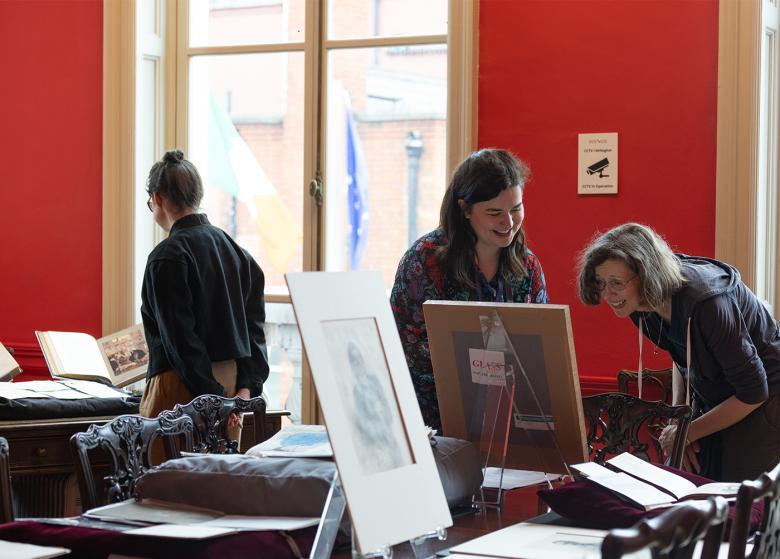 Three female figures in a red room, looking at a collection of drawings, books and other items that have been spread on a table. 