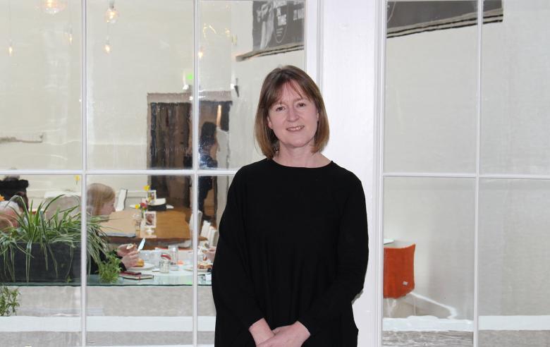 A female figure with a brown bob and a black dress stands in front of a large window