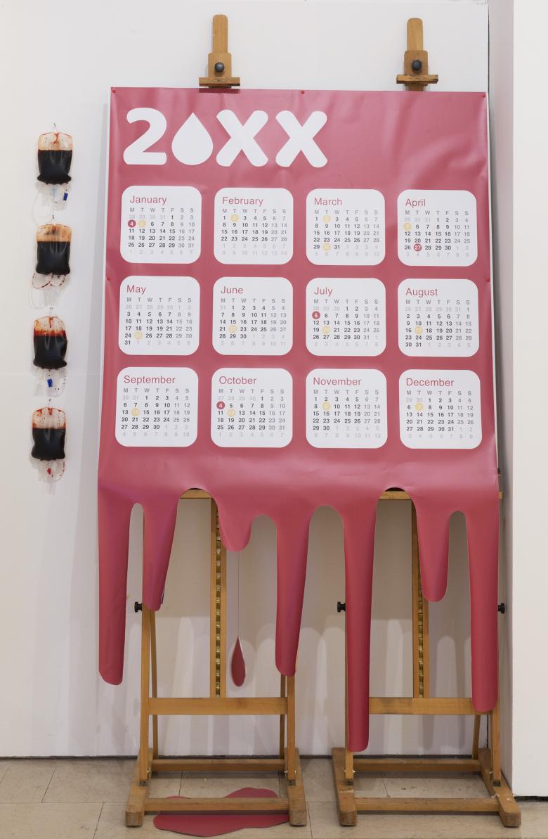 Installation shot of a large red calendar, mimicking blood dripping, mounted on an easel, with four blood bags hanging on the wall beside it