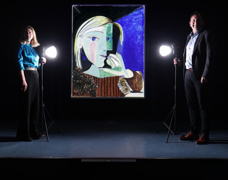Two figures stand in semi darkness on either side of a large painting by Picasso. In front of both of them is a spotlight.