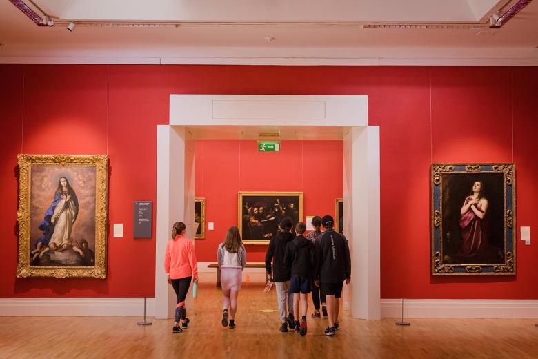 A group of young people walking away through an open doorway in an art gallery with red walls and gilt-framed oil paintings