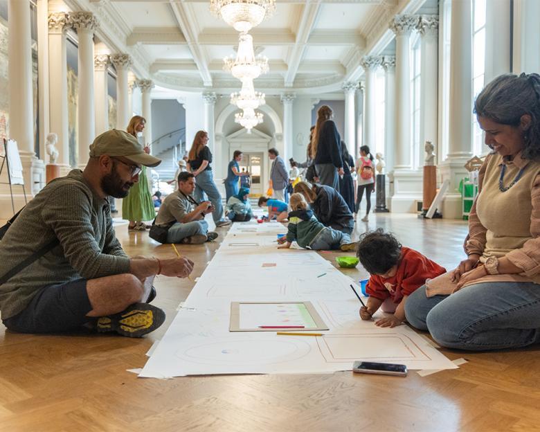 A family draw pictures on a long sheet of paper, which is laid on the floor of a grand room.