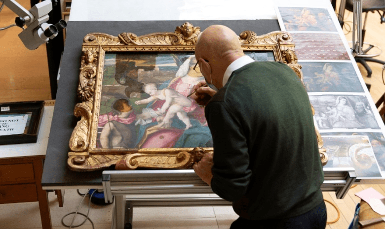 A man in a green jumper leans over an oil painting in a gilt frame with a small brush.
