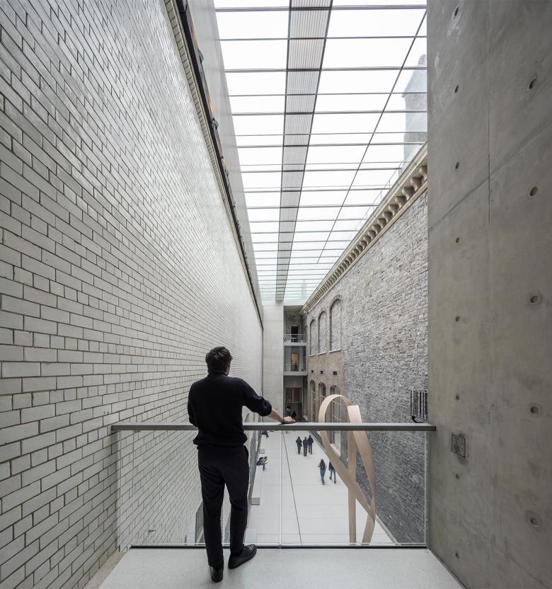 Photography of a male figure looking over a balcony railing to a grand internal courtyard made of grey stone.