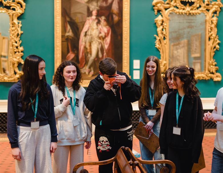 A teenage boy takes a photo on a bright orange camera in a grand gallery space. A group of five teenage girls watch on.