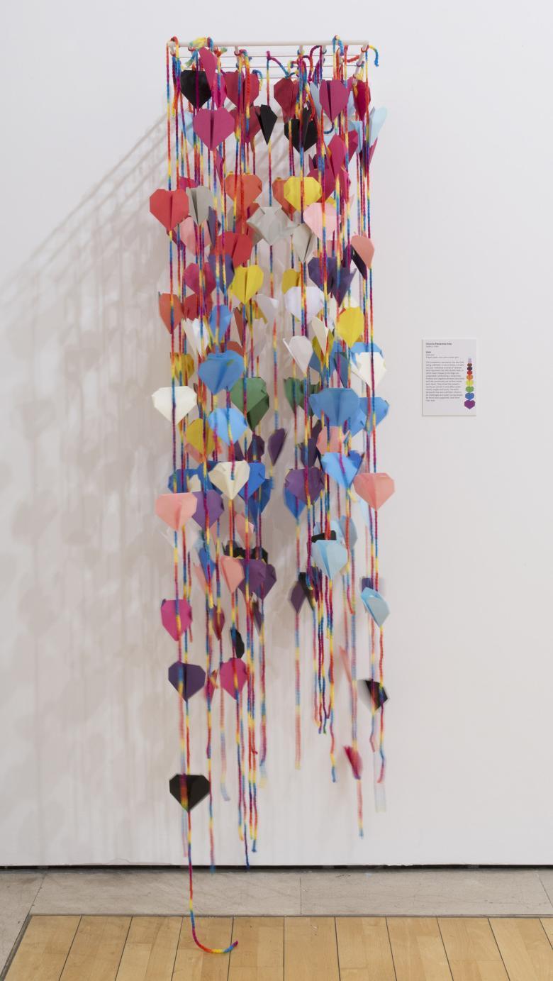 Installation shot of dozens of colourful origami hearts strung on strings and hanging down a white wall