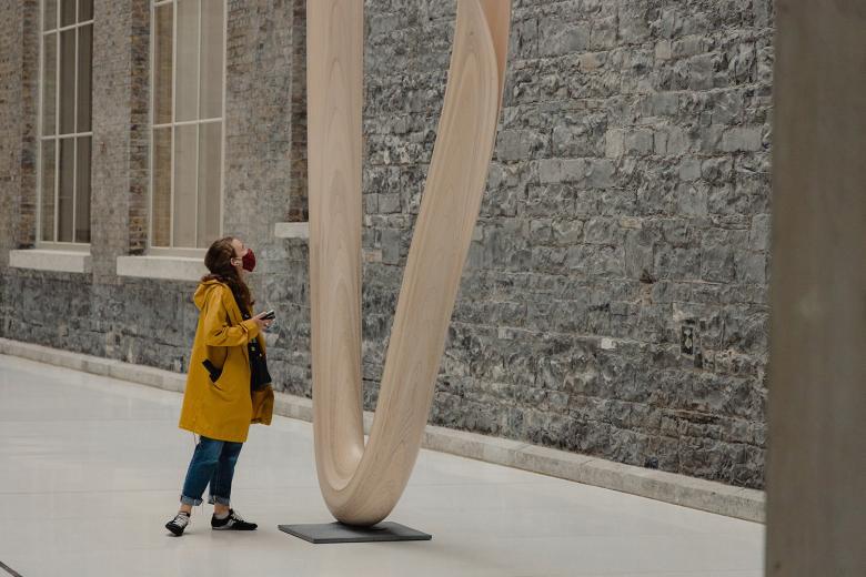Woman wearing a face mask looking at a large wooden sculpture in the Gallery