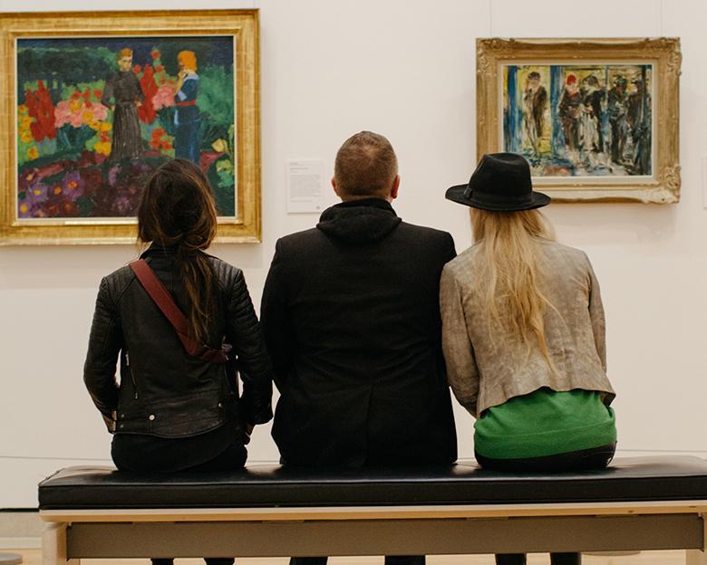 Photo of two women and a man seated on a bench, with backs to camera, looking at two paintings on a white wall.