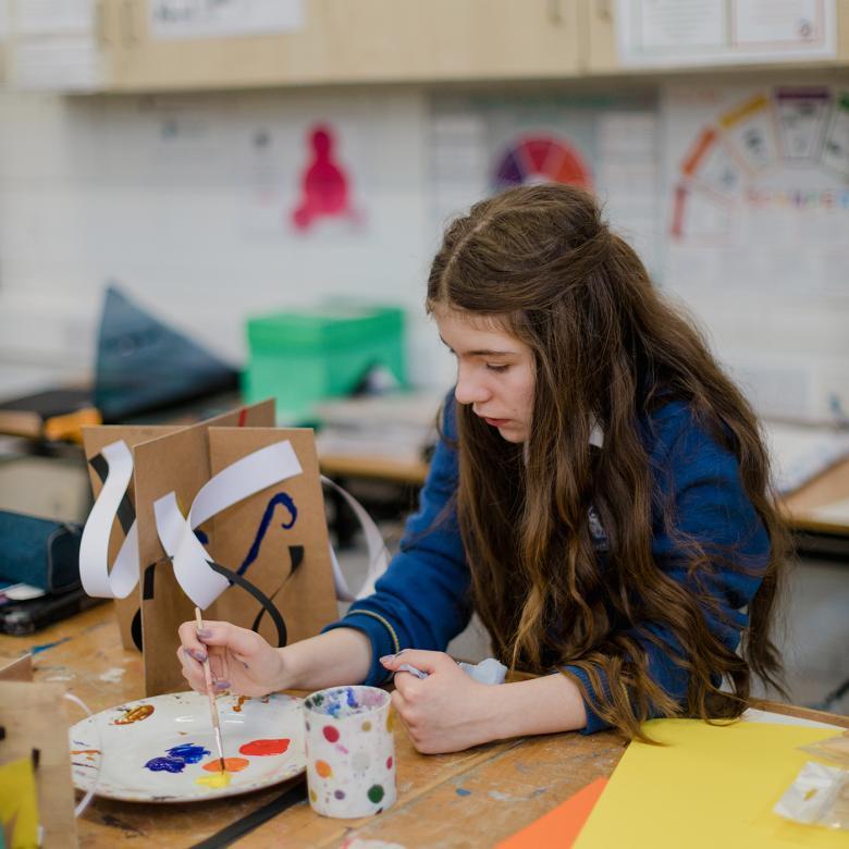 A young girl sits at a desk painting a cardboard and paper sculpture with brightly coloured paints