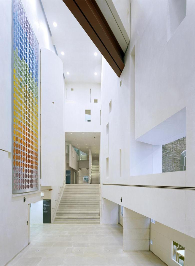 The large interior of a modern white building, with a large hanging tapestry on the left wall made up of a rainbow of spots.