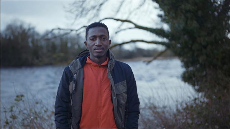 A still from a documentary film. A man in a red top and a jacket stands looking out, with a tree and field behind him