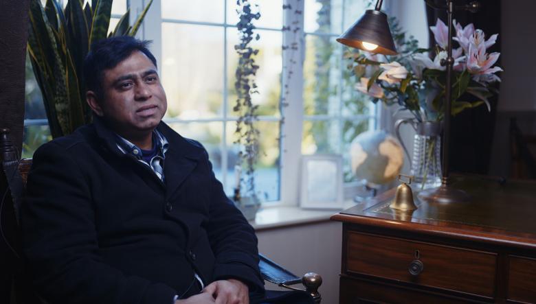 sits by a window, with his hands folded in his lap. 