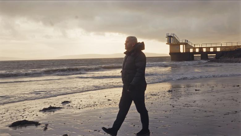 A still from a documentary film showing a figure walking across a beach a sunset.