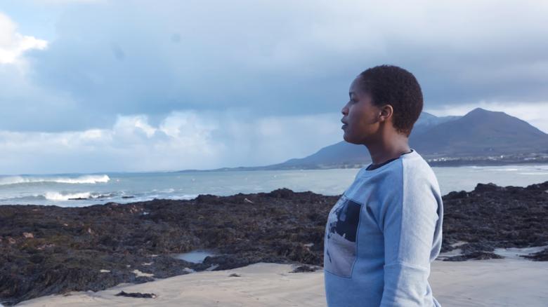 A still from a documentary film. A woman stands on a beach, looking out to the sea with a mountain in the background.