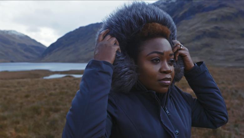A still from a documentary film. A woman wearing a winter coat pulls the hood up over her head. In the background, lakes and mountains