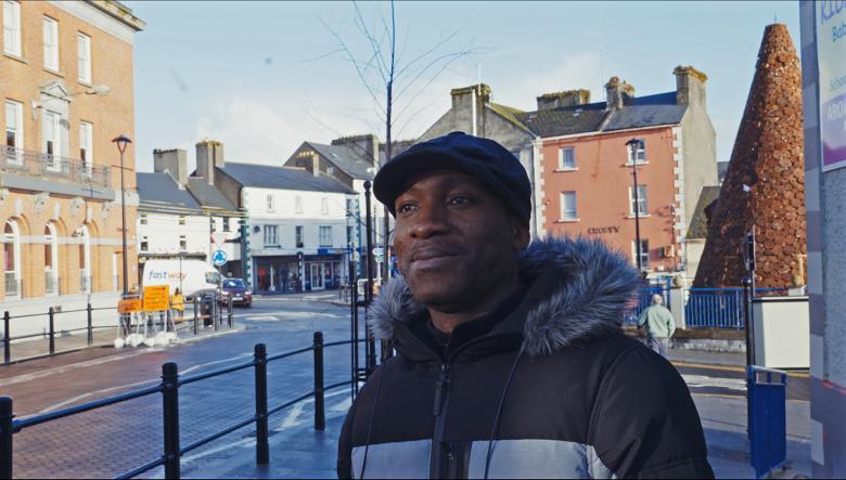 A still from a documentary film. A man wearing a flat cap and a coat with a fur-lined hood stands in a street