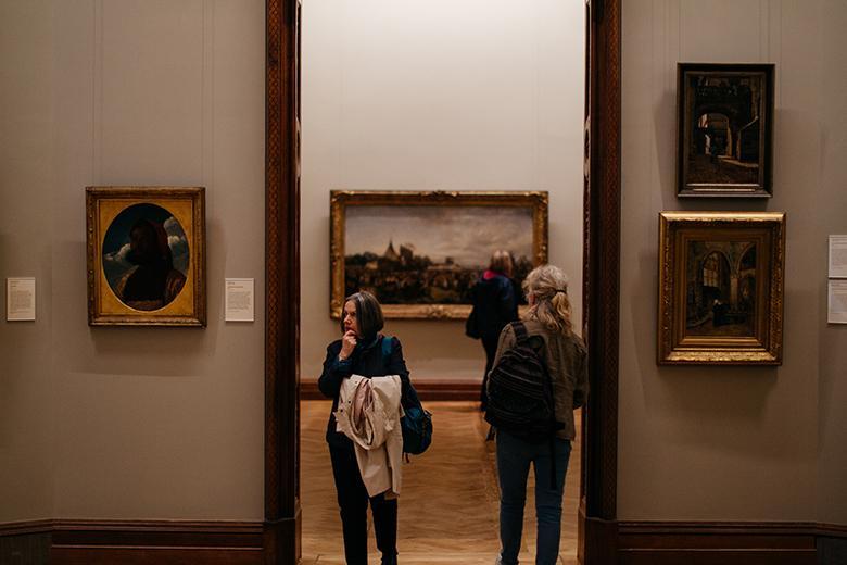 Two women passing each other in a doorway in a gallery room with gilt-framed paintings on the walls.