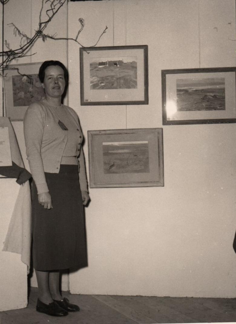 A woman stands beside a wall of framed artworks, looking at the camera. 