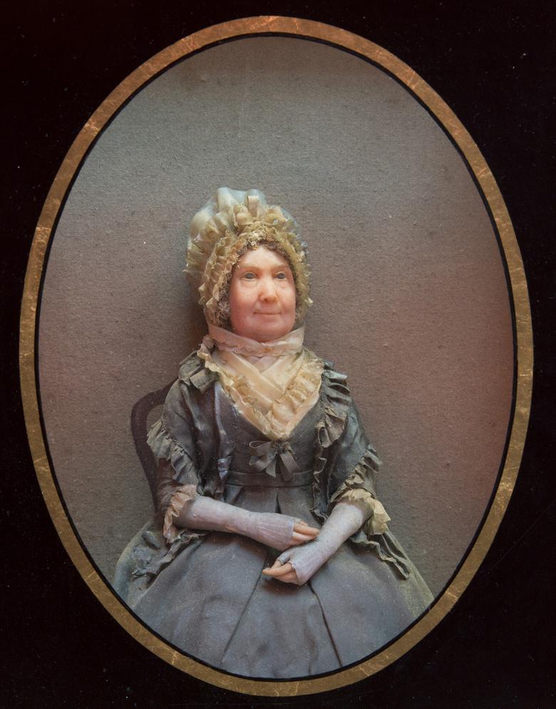 A wax figure of a woman seated with her hands in her lap. She is wearing a blue dress, and has a lace headdress on her head.