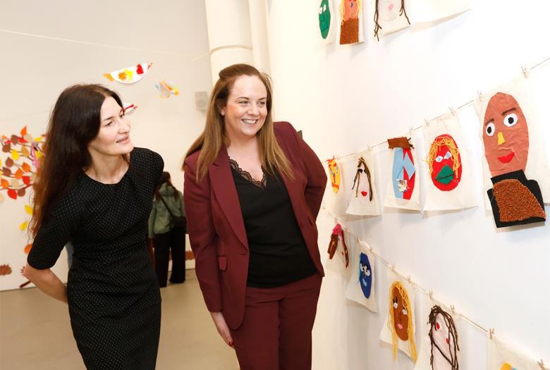 A female figure dressed in black stand beside a female figure wearing a brown suit, looking at artwork created by children.