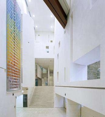 The large interior of a modern white building, with a large hanging tapestry on the left wall made up of a rainbow of spots.