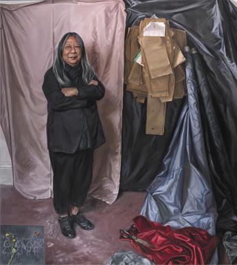 Painted full-length portrait of designer John Rocha wearing all black and standing in front of a wall covered in sheets of fabric and paper patterns.