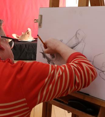 Man in a red and white striped top with grey hair drawing with pencil at an easel