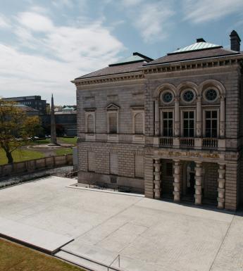 A colour photograph showing the stone facade of the Gallery's historic wing taken from a height. The sky is blue and streaked with white clouds.