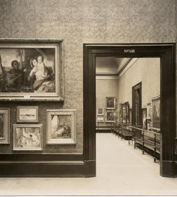 Black and white photo of paintings hung Salon-style on the wall with view through doorway into next gallery space