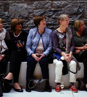 A group of people sit on a stone bench against an exposed stone wall. They appear to be listening intently to someone off-camera.