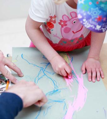 Photo of a young child drawing with an adult's hands stabilising the piece of paper.
