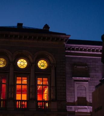 Nighttime photo of the exterior of the Merrion Square entrance to the National Gallery of Ireland with the windows illuminated and an outdoor sculpture silhouetted against the sky.