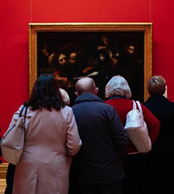 Photo of a group of people, with their backs to the camera, looking at Caravaggio's painting of the Taking of Christ, which is hanging on a red wall.