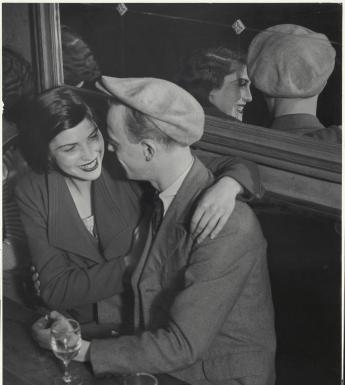 A black and white photograph of a woman and a man sitting side by side at a table, with two half full glasses of wine in front of them. They are turned towards each other, and the woman has her arm around the man's shoulder, and is smiling. Behind them is a large mirror in which we can see them reflected.