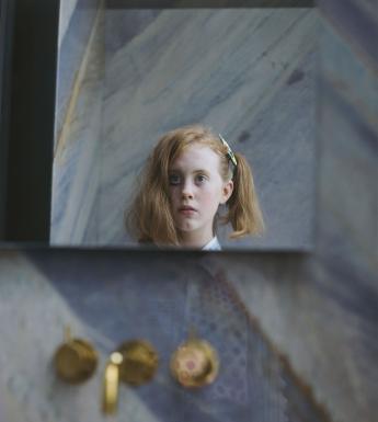 A photograph on paper. The head of a young girl is clearly visible, as though reflected in a square of mirror - on one side of her head her hair is pinned up in a ponytail, on the other side it is loose to her shoulders. One of her eyes has black markings under it. She is surrounded by abstract patterns - we can see her shirt faintly, as though reflected through a window. Out of focus beneath her face we see three brass fittings, like the fixtures of a bath.