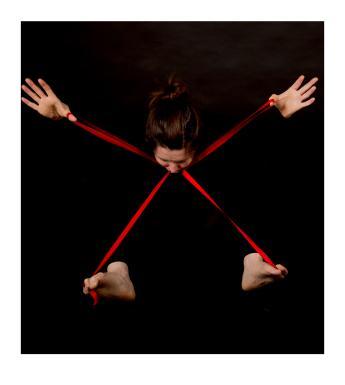 Emerging from a black background we see the head of a woman, eyes cast down, and her hands and feet. There is a red ribbon in the shape of an X connecting the part of her body we see.